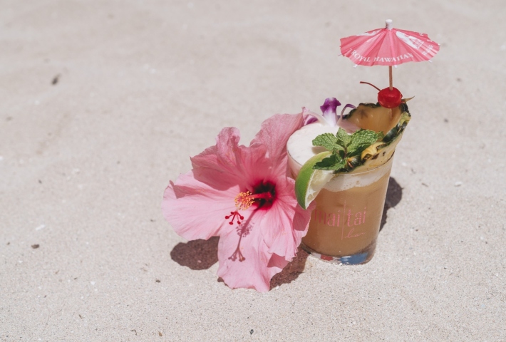 A Mai Tai bar cocktail and a pink hibiscus placed on the sand