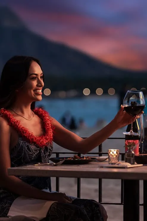 A woman raising a wine glass by the beach at sunset with Diamond Head in the background a romantic Royal Hawaiian dinner scene