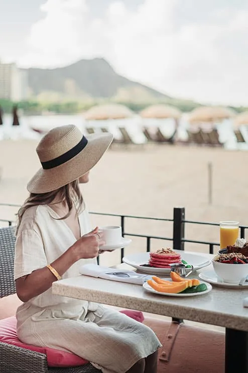 A woman enjoying breakfast on the Surf Lanai terrace overlooking Waikiki Beach, with pancakes, fruit, and coffee on the table, and Diamond Head and beach umbrellas in the background